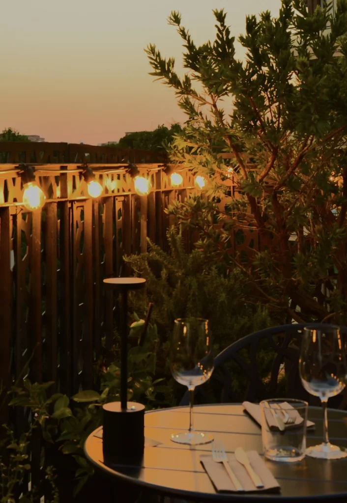 Nell'immagine, il primo piano su un tavolino apparecchiato presso il rooftop del Globus Hotel al tramonto, con due calici di vino, due bicchieri e una lampada da tavolo.
